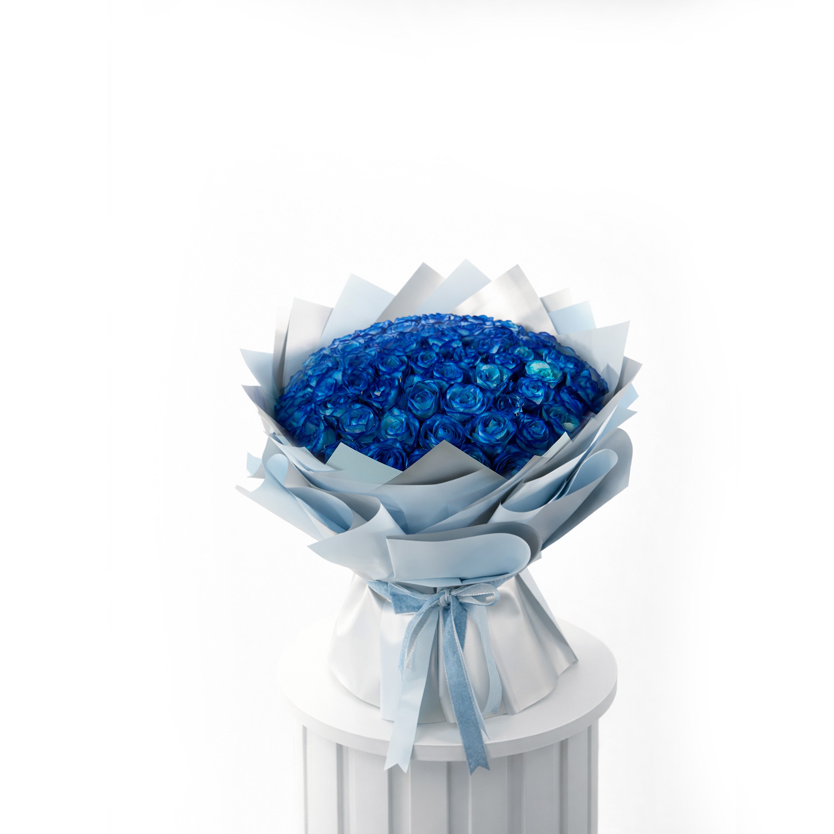 Bouquet of 50 blue roses wrapped in white paper with a blue ribbon on a white pedestal against a white background.