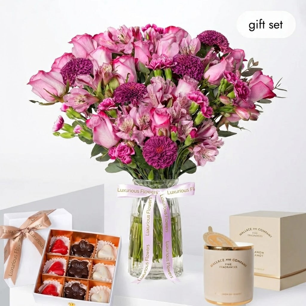 Bouquet of pink and purple flowers in a glass vase, surrounded by a box of chocolates, a candle, and a box of tea on a white background.