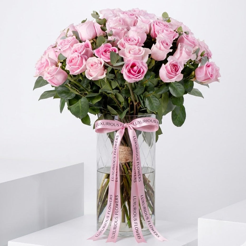 luxury bouquet of light pink roses with eucalyptus in a tall glass vase tied with a pink Luxurious Flowers ribbon, set against a clean white background.