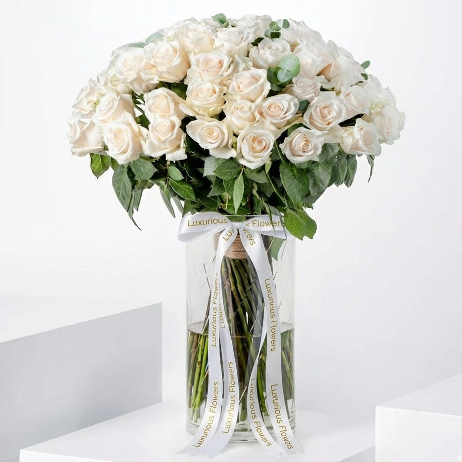 Bouquet of white roses in a clear vase with 'Luxurious Flowers' ribbon on a white background